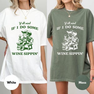 May include: White and moss green t-shirts with a graphic of a frog wearing a cowboy hat, holding a wine glass, and sitting on a bench. The text reads "Y'all mind IF I DO SOME WINE SIPPIN'" in green and white.