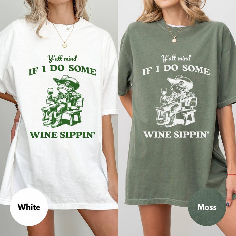 May include: White and moss green t-shirts with a graphic of a frog wearing a cowboy hat, holding a wine glass, and sitting on a bench. The text reads "Y'all mind IF I DO SOME WINE SIPPIN'" in green and white.