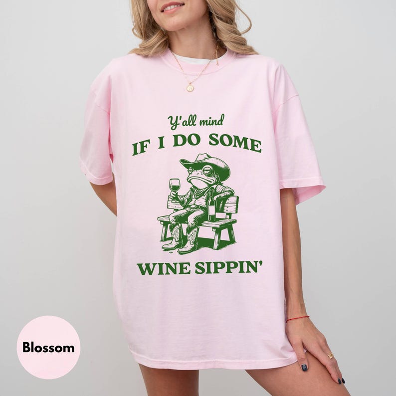 May include: A light pink t-shirt with a green graphic of a frog in a cowboy hat, boots, and holding a wine glass. The text reads "Y'all mind if I do some wine sippin'". The shirt is a casual, oversized fit.