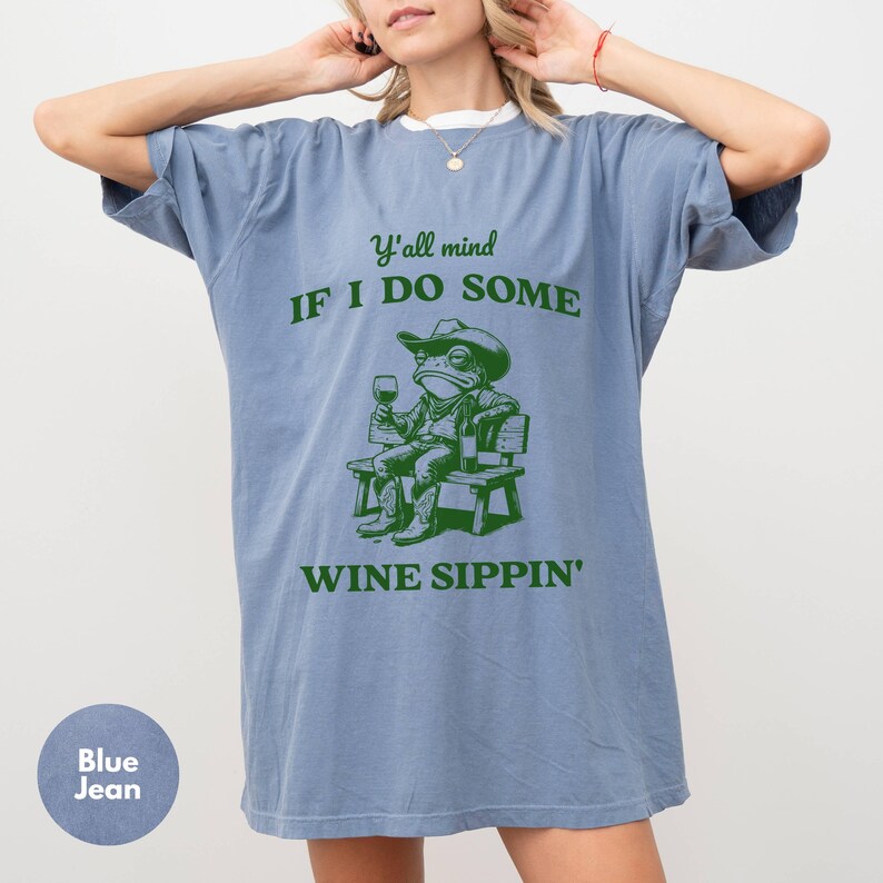 May include: Blue jean-colored t-shirt with a green graphic of a frog in a cowboy hat, boots, and a wine glass, seated on a bench. The text reads "Y'all mind if I do some wine sippin'."