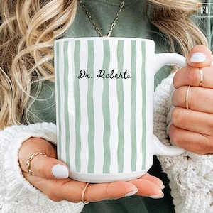 May include: White ceramic mug with vertical, uneven, light green stripes. The mug features the text "Dr. Roberts" in a handwritten style. The mug has a large handle and is held by a person.