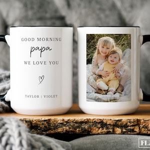 May include: White ceramic mug with a black handle and rim. One side features a photo of two children, the other side has the text "GOOD MORNING papa WE LOVE YOU" with a heart and names. The mug is on a wooden surface.
