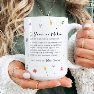 May include: White ceramic mug with the text "Difference Maker" and a definition, floral and heart accents. The mug is held by a person wearing a white sweater and gold rings. The mug is a standard size.
