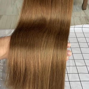 Puede incluir: Paquetes de extensiones de cabello castaño claro. El cabello es liso y tiene un brillo natural. Las extensiones se sujetan con pequeñas bandas de goma. El fondo es un patrón de cuadros blancos y negros.