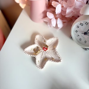 Peut inclure: Un vide-poche blanc en forme d'étoile avec une surface texturée et des éclats dorés. À l'intérieur se trouvent deux bagues dorées : une anneau simple et une autre ornée d'un cœur rouge avec un symbole d'œil blanc au centre.
