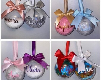 May include: Four sets of personalised Christmas baubles. Each set includes two clear glass baubles with different coloured ribbons and bows. The baubles are filled with white feathers and have names written on them in gold or silver lettering. The names are: Aurora, Sophia, Noah, Olivia, Luna, and Ava.