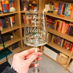 May include: A clear wine glass etched with the words "One Faerie Wine, Closer to Velaris". The glass has a long stem and a circular base. The background features a wooden bookshelf filled with books.