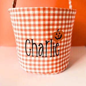 May include: A fabric bucket featuring an orange and white gingham pattern. The name Charlie is embroidered in black script lettering on the front, accompanied by an orange pumpkin applique with a black jack-o'-lantern face design.