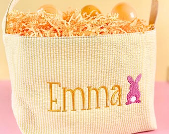 Puede incluir: Una cesta de Pascua a rayas amarillas y blancas con el nombre "Emma" bordado en dorado. Un conejito rosa está bordado junto al nombre. La cesta está llena de papel naranja y huevos de Pascua de plástico.