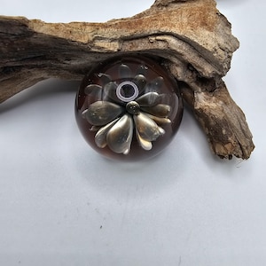 May include: A clear, round paperweight with a dark brown hue, featuring a silver-colored flower design. The flower has multiple petals and a detailed center. The paperweight is set against a white background and a piece of driftwood.