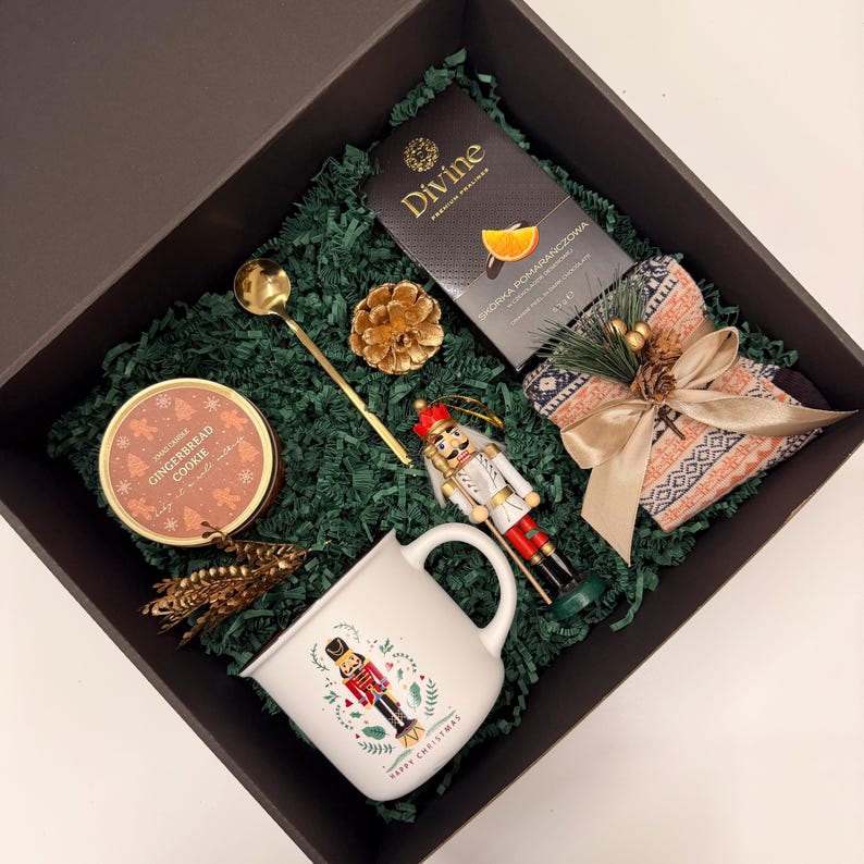 May include: A gift box filled with Christmas-themed items. Includes a white mug with a nutcracker design, a tin of gingerbread cookies, tea, a gold spoon, a nutcracker ornament, and patterned socks. The box is lined with green paper.