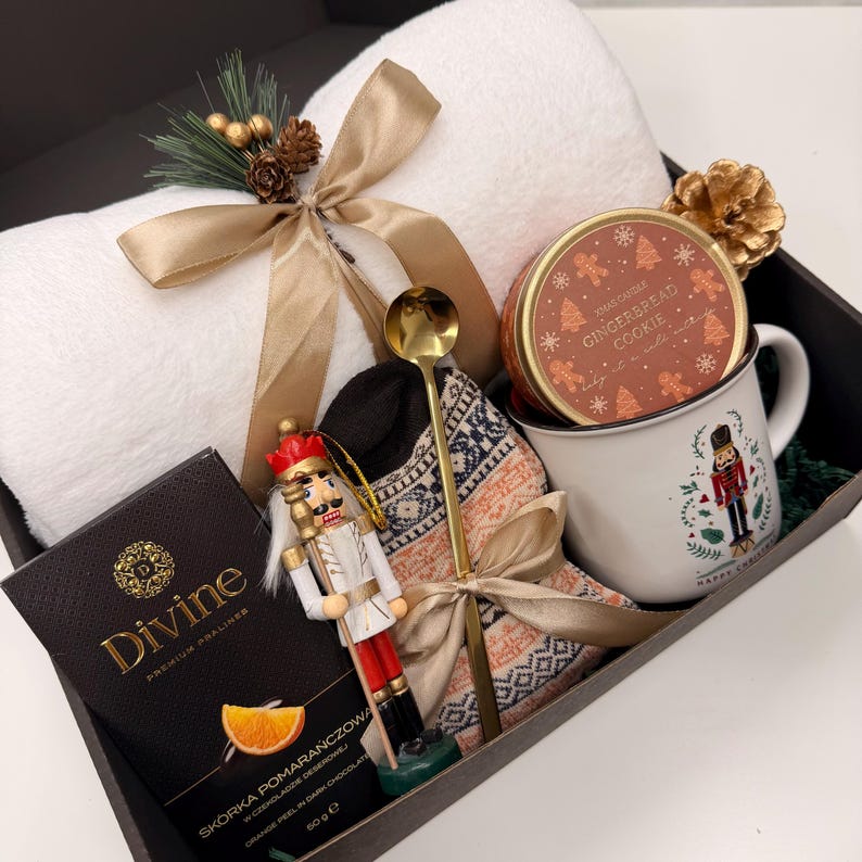 May include: A gift box filled with a white towel, a mug with a nutcracker design, a gingerbread cookie tin, a chocolate bar, a gold spoon, a decorative nutcracker, and a pair of patterned socks. The box is tied with a gold ribbon and decorated with pine and gold accents.