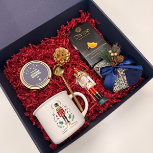 May include: A holiday gift box filled with red shredded paper, a white mug with a nutcracker design, a gold spoon, a nutcracker figurine, a candle, tea, and a small gift wrapped in blue ribbon.