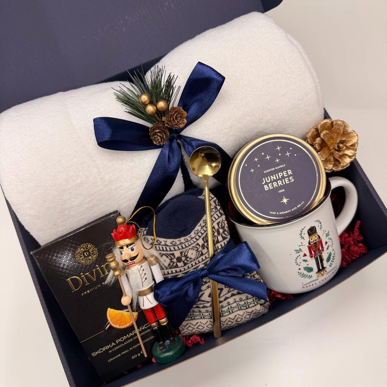 May include: A gift box with a white towel, a Juniper Berries candle, a mug with a nutcracker design, a nutcracker ornament, a gold spoon, a box of orange peel, and a pair of socks. The items are arranged in a navy blue box.