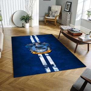 May include: A rectangular blue rug with white stripes and a Ravenclaw crest. The crest features a bird emblem and the word "Ravenclaw" in gold. The rug is placed on a wooden floor in a living room setting.