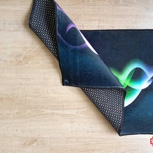 May include: A rectangular rug with a dark blue background and a colorful abstract design in shades of purple, green, and white. The rug's underside is black with a white dotted pattern. The rug is partially folded, revealing the underside.