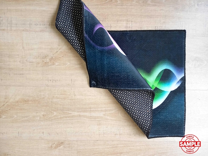 May include: A rectangular rug with a dark blue background and a colorful abstract design in shades of purple, green, and white. The rug's underside is black with a white dotted pattern. The rug is partially folded, revealing the underside.
