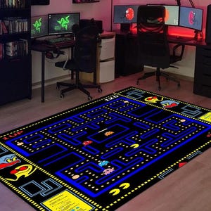May include: A rectangular black rug featuring a Pac-Man game design. The rug has a blue maze pattern with colorful Pac-Man characters and ghosts. The border is yellow with game-related graphics and text. The rug is placed in a gaming room.