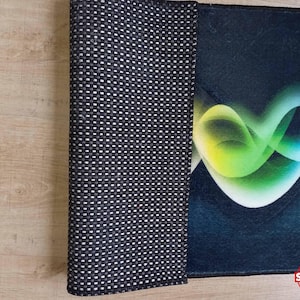 May include: A rolled-up rug with a black and white dotted underside and a colorful abstract design on the top. The design features flowing lines of green, yellow, and blue against a dark blue background. The rug is on a light brown wooden surface.