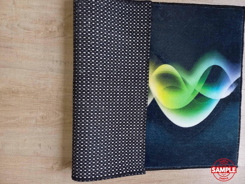 May include: A rolled-up rug with a black and white dotted underside and a colorful abstract design on the top. The design features flowing lines of green, yellow, and blue against a dark blue background. The rug is on a light brown wooden surface.