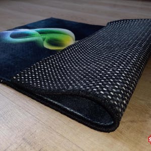 May include: A rectangular rug with a dark blue surface and a colorful, abstract design. The rug's underside is black with a textured, non-slip pattern. The rug is partially rolled up, revealing the underside.