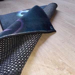 May include: A rolled-up, dark blue rug with a black border and a non-slip, checkered underside. The top surface features a design with blue, purple, and green hues. The rug is partially unrolled, revealing its texture and pattern.
