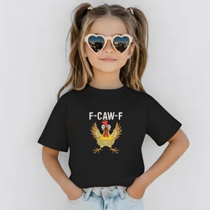 K&ouml;nnte beinhalten: Ein schwarzes T-Shirt mit dem Text "F-CAW-F" und einem Cartoon-H&uuml;hner-Design. Das Huhn ist gelb, rot und braun. Die Person auf dem Bild tr&auml;gt herzf&ouml;rmige Sonnenbrillen.