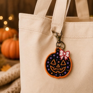 May include: A beige canvas tote bag featuring a hanging pumpkin-shaped keychain. The keychain is black with orange trim, decorated with small purple and orange stars, a carved jack-o'-lantern face, and a small white and red bow at the top.