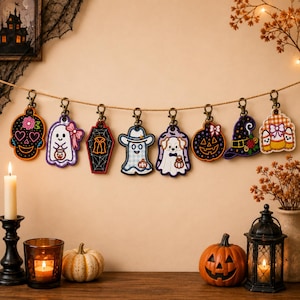 May include: A string of eight embroidered Halloween-themed keychains hanging from a twine rope. The designs include a sugar skull, a ghost with a pink bow, a coffin, a ghost in a cowboy hat, a dog in a ghost costume, a jack-o'-lantern, a witch hat, and candy corn.