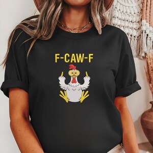 K&ouml;nnte beinhalten: Schwarzes T-Shirt mit dem gelben Text "F-CAW-F". Das Shirt zeigt eine Cartoon-H&uuml;hner-Illustration, die den Mittelfinger zeigt. Das Huhn ist wei&szlig; mit gelben F&uuml;&szlig;en und einem roten Kamm.