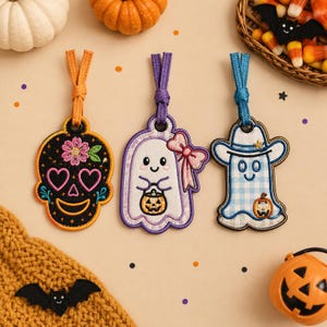May include: Three embroidered hanging ornaments featuring a black sugar skull with pink heart eyes, a white ghost with a pink bow holding a pumpkin bucket, and a blue gingham ghost wearing a cowboy hat and holding a small pumpkin.