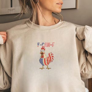 K&ouml;nnte beinhalten: Beiges Sweatshirt mit einem Cartoon-Hahn in Rot, Wei&szlig; und Blau, &auml;hnlich der amerikanischen Flagge. Der Text "F-CK-E-F" befindet sich &uuml;ber dem Hahn. Ein bequemes, l&auml;ssiges Kleidungsst&uuml;ck.