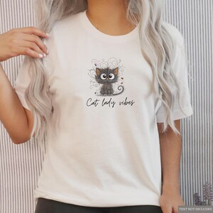 以下が含まれることがあります： 白いTシャツ。漫画の猫のイラストと「Cat lady vibes」の文字がプリントされています。猫はグレーで大きな目を持ち、装飾的な渦巻きと小さなカラフルなアクセントに囲まれています。Tシャツは半袖です。