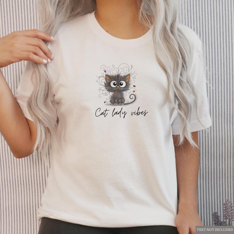 以下が含まれることがあります： 白いTシャツ。漫画の猫のイラストと「Cat lady vibes」の文字がプリントされています。猫はグレーで大きな目を持ち、装飾的な渦巻きと小さなカラフルなアクセントに囲まれています。Tシャツは半袖です。