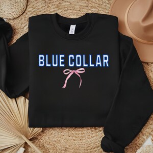 May include: Black crewneck sweatshirt with the text "BLUE COLLAR" in blue and white, and a pink bow graphic. The sweatshirt is displayed on a neutral woven surface with a hat and decorative elements.