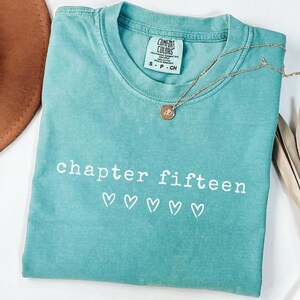 May include: A turquoise Comfort Colors t-shirt with the words "chapter fifteen" and five heart symbols printed in white. A gold necklace with a pendant rests on the shirt. The shirt is folded on a white surface.