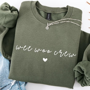 May include: An olive green sweatshirt with the text "wee woo crew" in white script and a white heart. A silver necklace with a circular pendant is draped on the sweatshirt. The sweatshirt is folded on top of a pair of blue jeans.