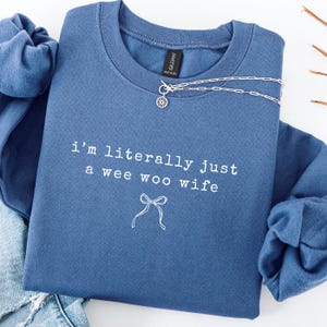 May include: A blue sweatshirt with the text "i'm literally just a wee woo wife" in white. A silver necklace with a small pendant is draped across the sweatshirt. The sweatshirt is folded on top of a pair of blue jeans.