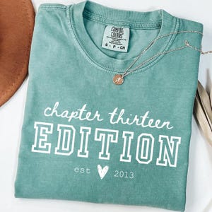 May include: A teal Comfort Colors t-shirt with the words "chapter thirteen EDITION" printed in white. Below the text is "est" a heart symbol, and "2013". A gold necklace with a pendant rests on the shirt.