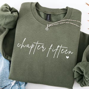 May include: An olive green sweatshirt with the words "chapter fifteen" in white script and a small white heart. A silver necklace with a sunburst pendant is draped on the sweatshirt. The sweatshirt is folded with a pair of blue jeans.
