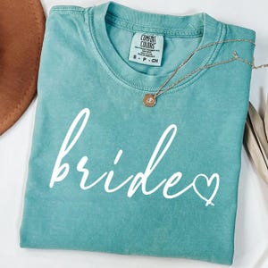 May include: A teal Comfort Colors t-shirt with the word "bride" in white script and a heart. A gold necklace with a pendant rests on the shirt. A brown hat is partially visible in the upper left corner.