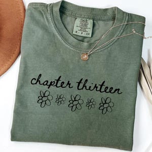 May include: Sage green t-shirt with the words "chapter thirteen" and flower graphics in black. A silver necklace with a small pendant rests on the shirt. The shirt is folded and has a tag that says "Comfort Colors".