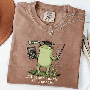 May include: Brown Comfort Colors t-shirt with a cartoon frog wearing a mortarboard and holding a calculator and pointer. The shirt reads "I'll teach math 'til I croak." A silver necklace with a pendant is also visible.