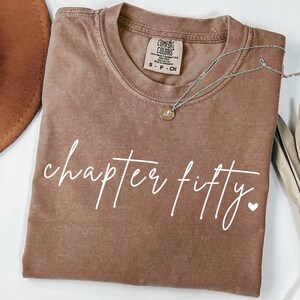 May include: A brown short-sleeve t-shirt with a crew neckline and a Comfort Colors brand label at the inner collar. The front features the white cursive text "chapter fifty." followed by a small heart symbol. A silver necklace rests on the fabric.