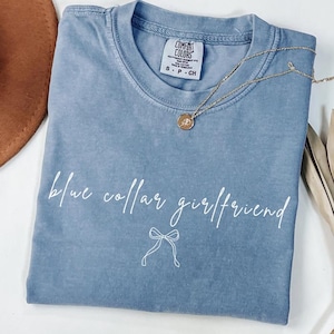 May include: A blue Comfort Colors t-shirt with the phrase "blue collar girlfriend" in white script. A gold necklace with a pendant and a brown hat are also visible.