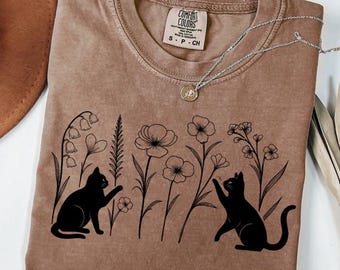 May include: A brown Comfort Colors t-shirt featuring a black graphic of two cats among flowers. The shirt has a crew neck and short sleeves. A silver necklace with a pendant is draped across the shirt. The shirt has a tag that reads "Comfort Colors".