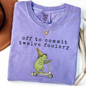 May include: A light purple t-shirt with a graphic of a green frog wearing a party hat and riding a scooter. The text above the frog reads, "off to commit twelve foolery." The shirt features a Comfort Colors brand label at the inner neckline.