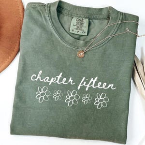 May include: An olive green t-shirt with the words "chapter fifteen" in white script and white flower graphics. A gold necklace with a small pendant rests on the shirt. The shirt has a Comfort Colors tag.