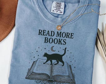 May include: A light blue t-shirt with the text "READ MORE BOOKS" and a graphic of a black cat walking on an open book under a crescent moon and stars. A gold necklace with a pendant is on the shirt.