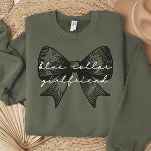 May include: Olive green sweatshirt with a black bow graphic and the words "blue collar girlfriend" in white script. The sweatshirt has a crew neck and long sleeves. A casual, comfortable top.